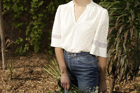 A model stands in a garden wearing a loose-fitting white shirt. She wears it tucked into high-waisted jeans. The sleeves are gathered at the top and flare out a little. They are elbow length and stop level with the model&#39;s waist.