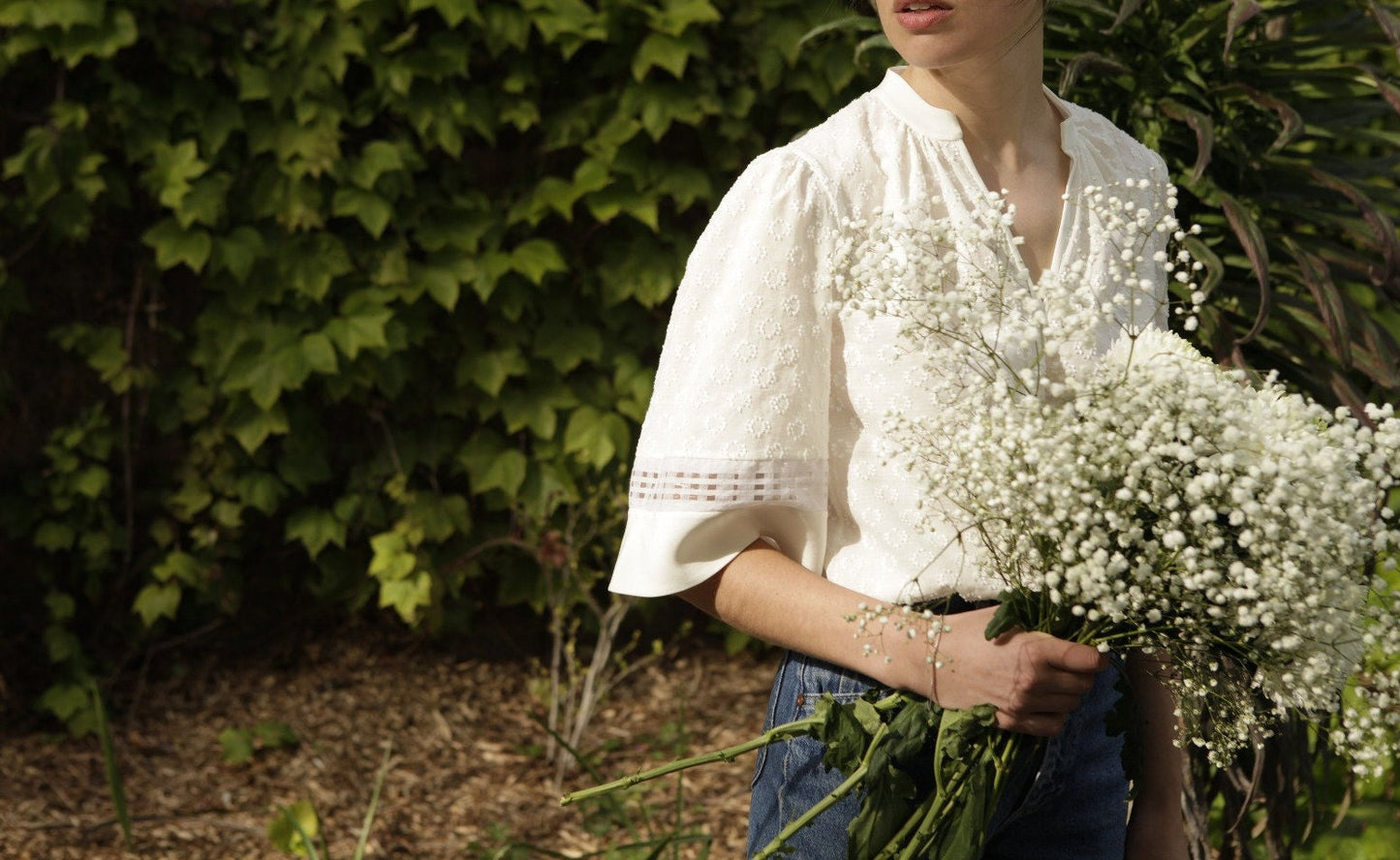 The model holds a bouquet of white flowers and looks over her shoulder. Her sleeves are made from 3 horizontal stripes of fabrics. The main sleeve is textured cotton, then a thin stripe of checked organza and a wider stripe of silk crepe at the hem.