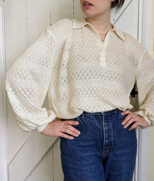 A model stands with her hands on her hips wearing a lace textured polo shirt loosely tucked into high-waisted jeans. The voluminous sleeves billow out over her cuffs. Her cream shirt has a short placket which she wears with a couple of buttons open.