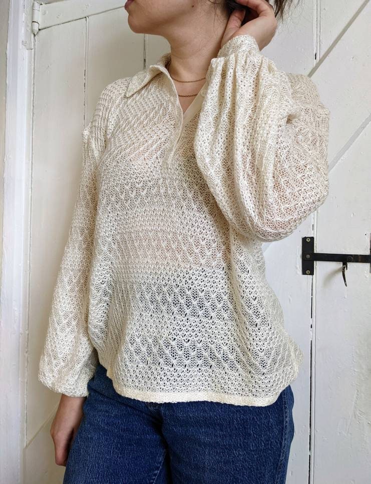 A closeup of the polo shirt from the front. The model raises one hand to her hair to show the fabric gathered at the cuff. She wairs the top over high waisted jeans and you can just see the blue denim through the slightly sheer fabric of the top.