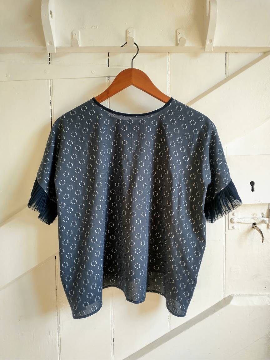 This is the back of the Frida top, shown hanging on a wooden hanger from a hook on a wooden farmhouse door. The top is cut as a square so it drapes from the shoulders and falls slightly longer at the sides. The neckline is a little higher at the back