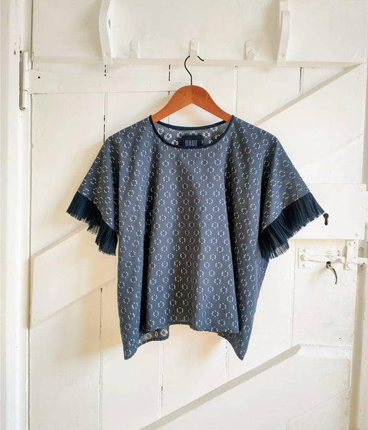 A dusky blue top hangs against a white wooden door. It is a square boxy shape with short t-shirt sleeves. The sleeves and neck trim are navy blue, as is the brand label on the inside of the back neck.