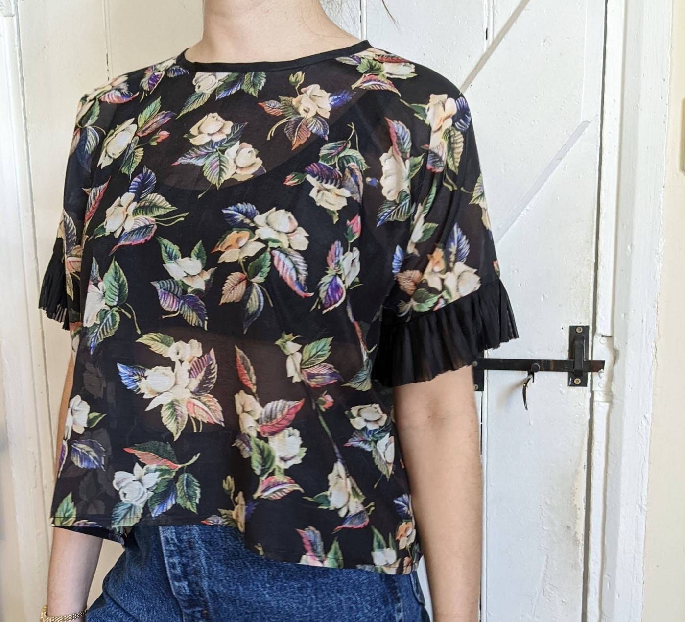 A model wears a loose-fitting floral patterned top. The top falls to above her hips and just covers the top of her high-waisted jeans. The hem of the sleeve sits a couple of inches above her elbow. A rounded neckline sits above her collarbones.