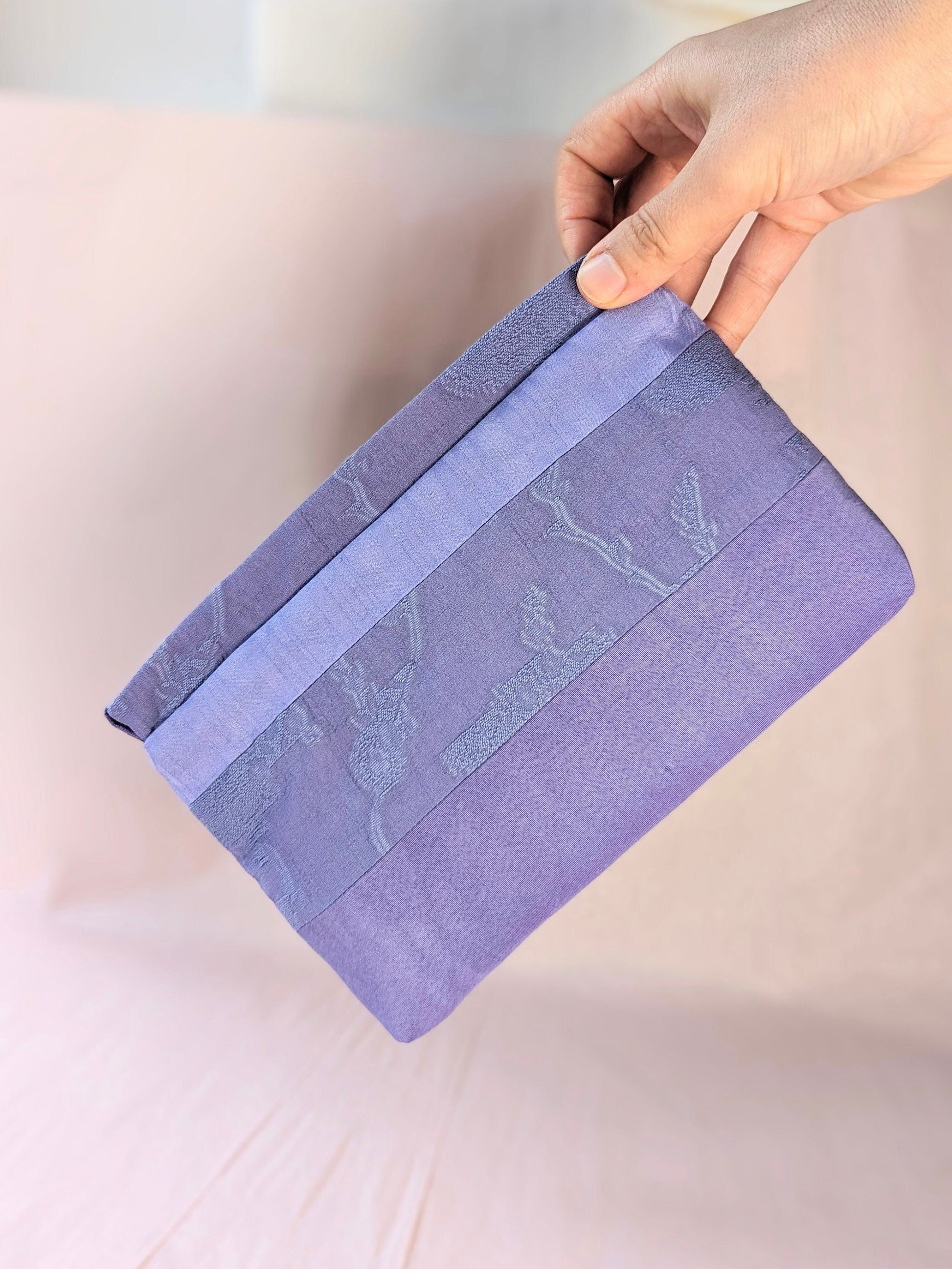 Image shows a white hand holding a lavender coloured, rectangle shaped purse. It is made of unequal stripes of fabric. Each fabric is a similar shade of purple but each is a different texture. The background is pale pink.